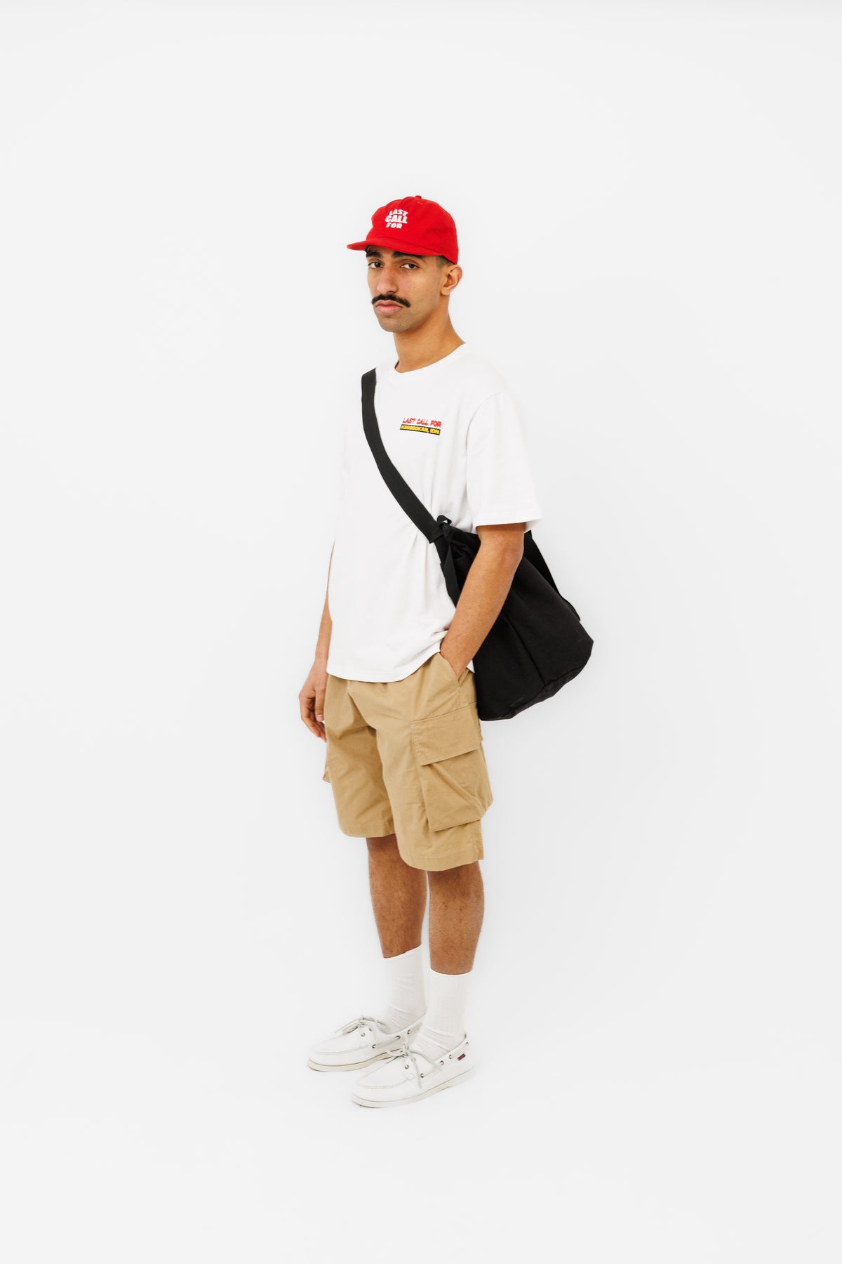Homme portant le Lombok Short beige Last Call For avec un t-shirt blanc, une casquette rouge et un sac bandoulière noir.