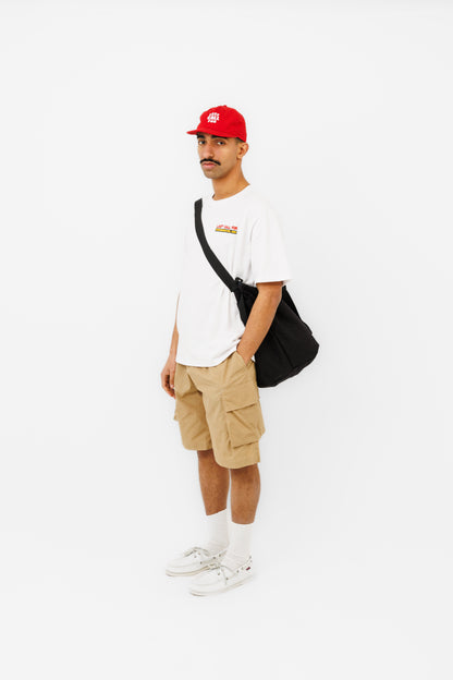 Homme portant le Lombok Short beige Last Call For avec un t-shirt blanc, une casquette rouge et un sac bandoulière noir.